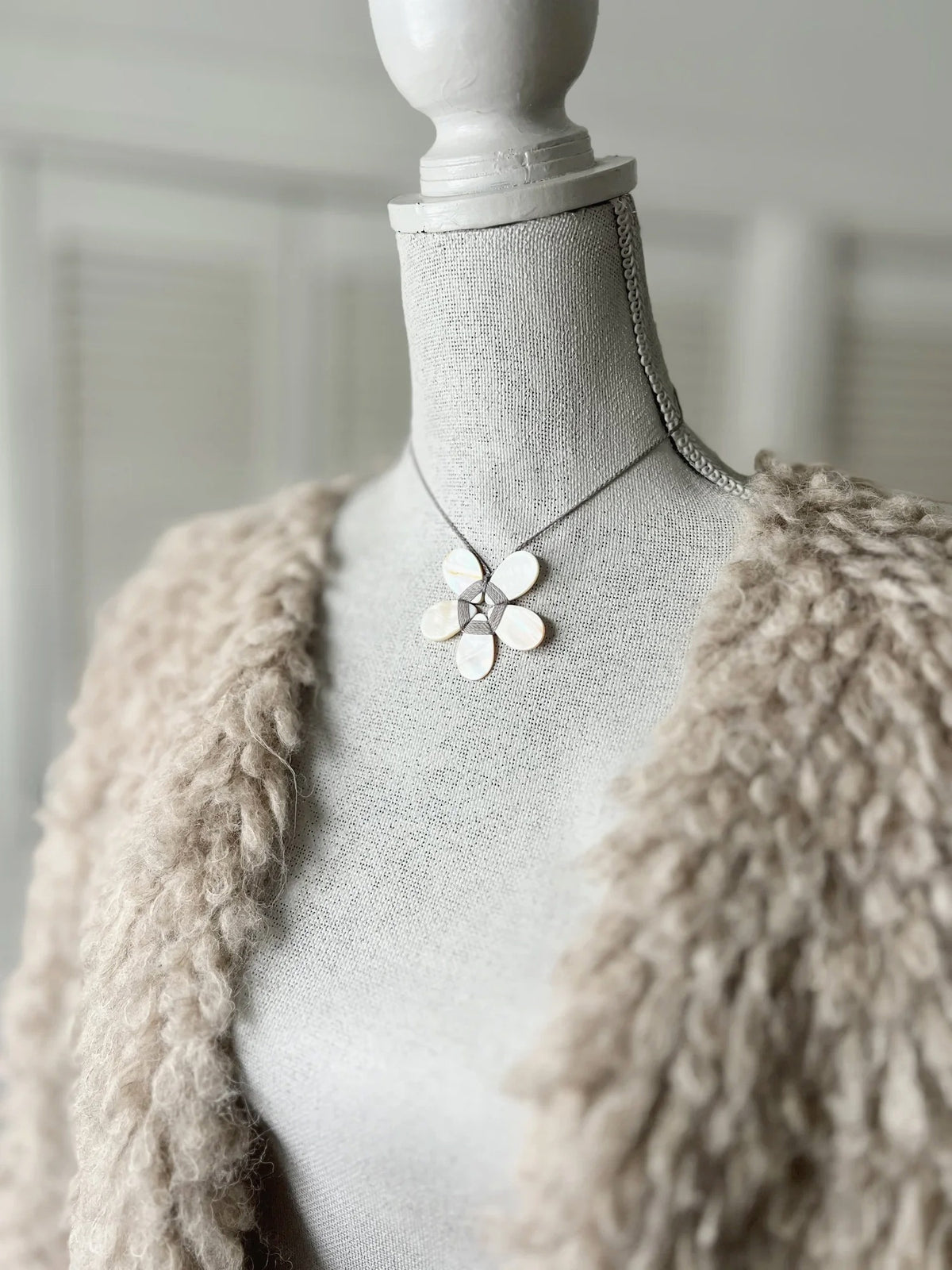 A cream-colored mannequin showcases a textured beige jacket paired with the ANN LIGHTFOOT - FLAT MOTHER OF PEARL FLOWER NECKLACE, highlighting a striking white-petaled flower pendant with a metallic center suspended from an adjustable braided cord. The background is softly blurred.