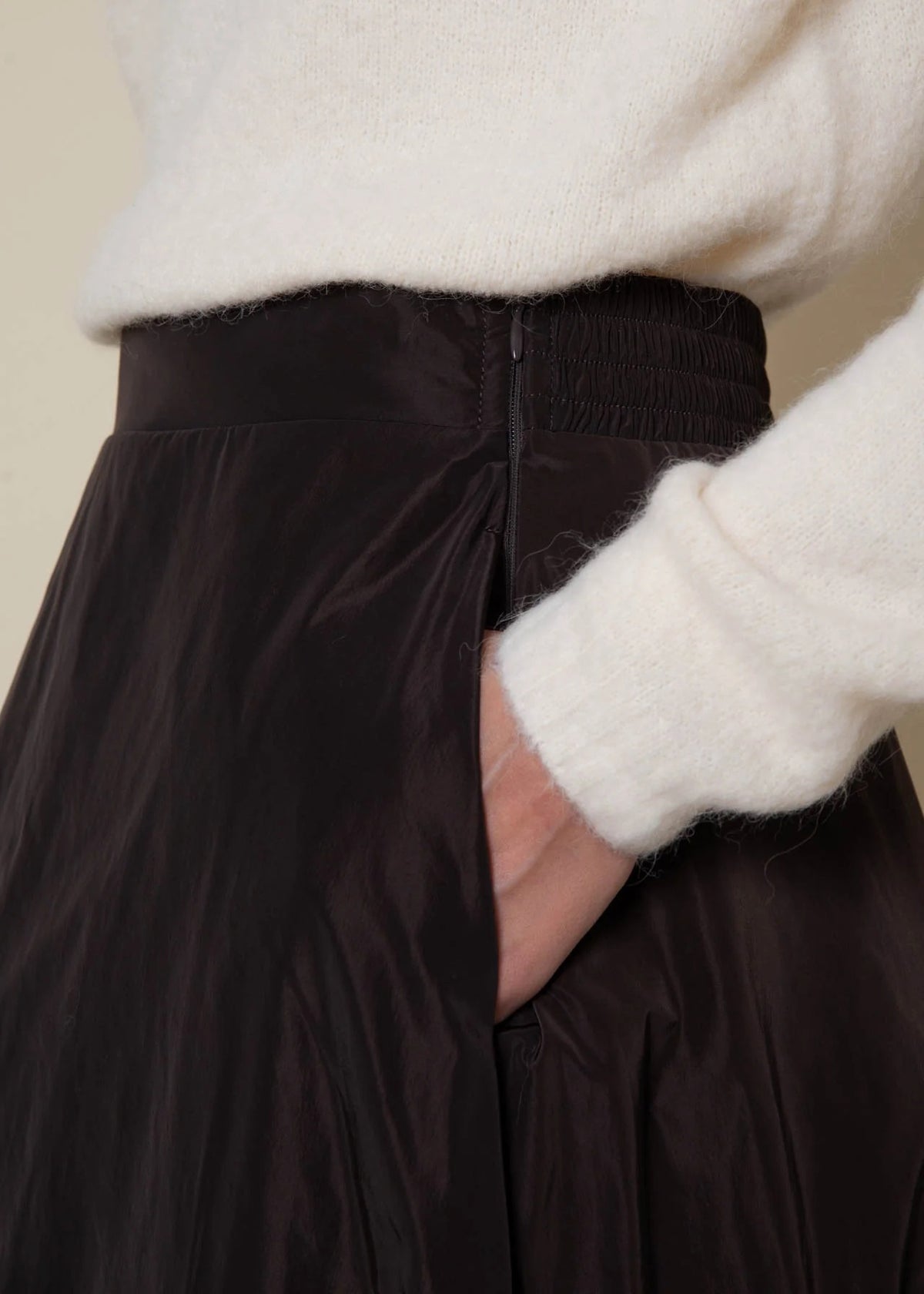 A person wearing a white sweater has their hand in the pocket of the VICARIO CINQUE GRANADA Taffeta Skirt in Marron Brown, a high-waisted skirt crafted from memory taffeta with a subtle sheen.