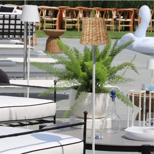 A modern poolside area features white lounge chairs, a potted fern, EMPIRE Water Hyacinth Woven Wicker Shade XXL by Zafferano America, white swan float, and a cordless lamp with glass water bottle on the table.