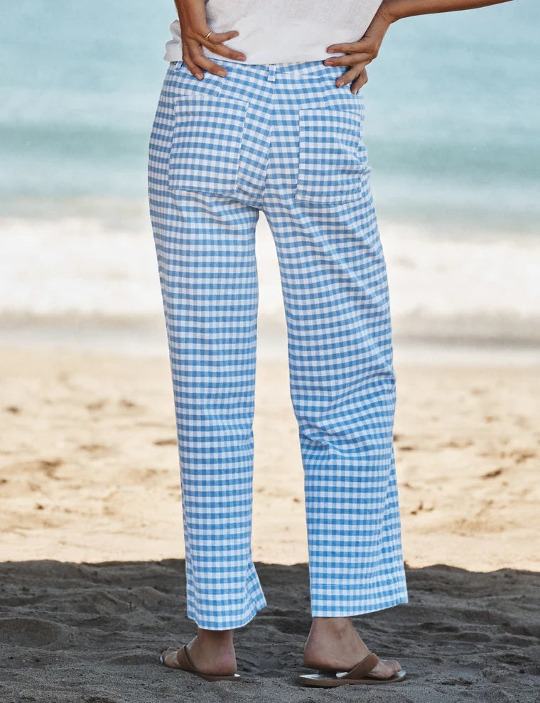 A person stands on a sandy beach wearing the FRANK & EILEEN WEXFORD Wide Leg Linen Pant in Blue Gingham, paired with a white shirt and beige sandals. The ocean is visible in the background.