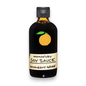 A brown bottle with a black cap, labeled “MOMOFUKU - SOY SAUCE,” featuring an orange peach graphic, delivers premium restaurant-grade soy sauce from the MOMOFUKU brand for exceptional flavor.