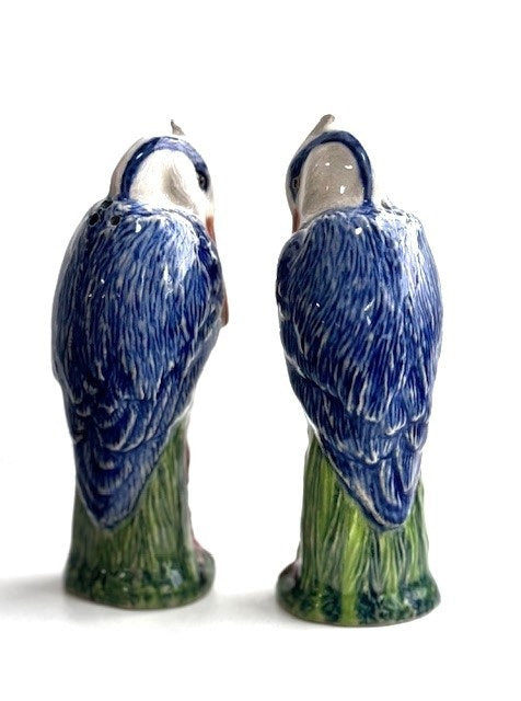 The QUAIL DESIGNS QUAIL CERAMICS Heron Salt and Pepper Shakers feature two hand-painted blue and white herons on green grass, facing each other. Dishwasher safe for easy cleaning.