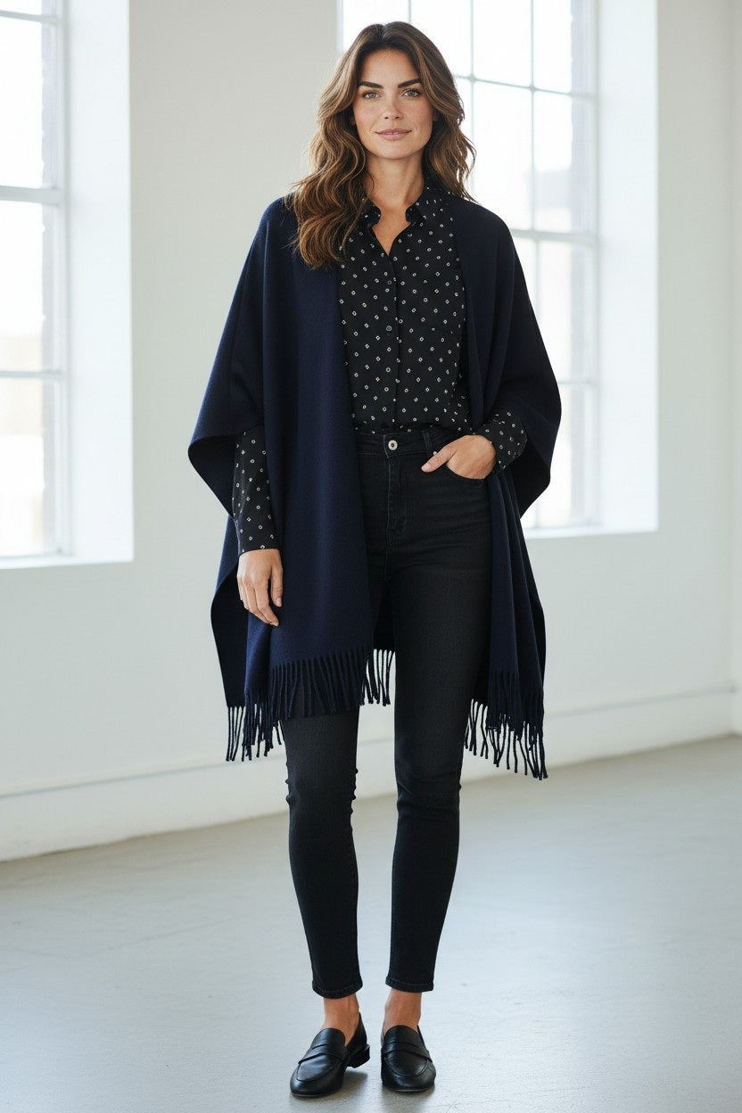 A woman stands indoors wearing the HARTFORD - VIRGINIE WOOL CAPE by HARTFORD over a black polka dot shirt, black jeans, and black loafers. She smiles softly with her hands relaxed at her sides, while bright windows illuminate the background.