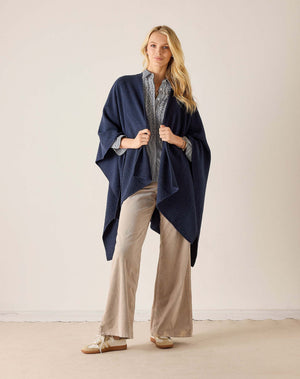 A woman stands indoors wearing the MERSEA - Modern Travel Wrap in Slate draped over a light gray patterned shirt, beige wide-leg pants, and tan shoes. She looks at the camera with a relaxed expression and long blonde hair.