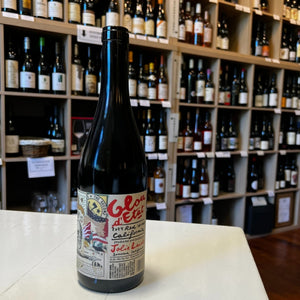 A bottle of JOLIE-LAIDE ’GLOU D’ETAT’ 2024 from WINE: KELLOGG SELECTIONS, featuring a colorful label, sits on a white table in front of shelves stocked with assorted wine bottles in a store.