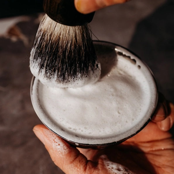 The BAUDELAIRE - M.B. SHAVING SET by BAUDELAIRE includes a luxurious shaving brush and premium shaving soap, presented in an elegant box featuring a delightful hat illustration and “shaving set” printed on the front. Displayed outdoors atop lush moss, this refined ensemble adds classic sophistication to your grooming routine.