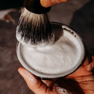 The BAUDELAIRE - M.B. SHAVING SET by BAUDELAIRE includes a luxurious shaving brush and premium shaving soap, presented in an elegant box featuring a delightful hat illustration and “shaving set” printed on the front. Displayed outdoors atop lush moss, this refined ensemble adds classic sophistication to your grooming routine.