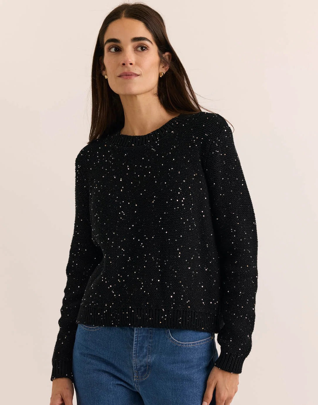 A woman with long dark hair, dressed in the HINSON WU - QUEENIE LONG SLEEVE SEQUIN SWEATER IN BLACK by HINSON WU and blue jeans, stands against a plain light background, smiling gently at the camera.