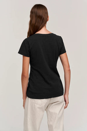 A person with shoulder-length brown hair is standing with their back to the camera. They are wearing a VELVET BY GRAHAM & SPENCER's LILITH V NECK SLUB COTTON TEE in blue and light blue jeans. The background is a plain, light-colored wall.