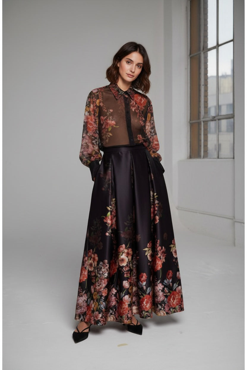 A woman wears a sheer, floral blouse paired with the MILVA MI - LONG MAXI SKIRT by MILVA—a shiny, high-waist skirt featuring a colorful floral print—and pointed black heels, posing against a simple gray studio backdrop.
