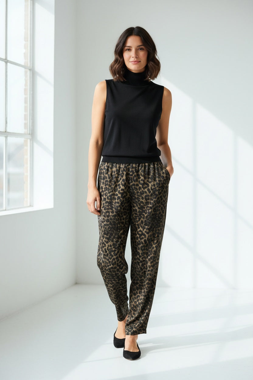 A woman with long brown hair wears the HINSON WU ASHLEY Sleeveless Turtleneck Knit Tank in black, paired with leopard print pants, standing against a plain light background with one hand on her hip.