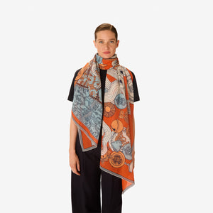 A woman with light skin and brown hair tied back wears a black outfit and the INOUI Editons Octopus Scarf in orange by INOUI, featuring intricate hand-printed patterns in orange, blue, and white draped over her shoulders.