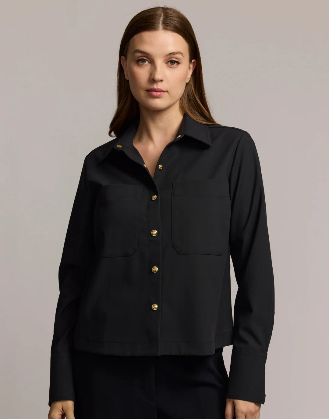 A woman with long brown hair models the HINSON WU - ASTRID LONG SLEEVE TRAVELER&#39;S TWILL SHIRT JACKET in dark green over an olive top and blue jeans, showcasing versatile HINSON WU travel style with gold hoop earrings against a neutral background.