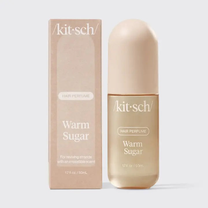 A bottle of KITSCH - HAIR PERFUME MIST IN WARM SUGAR stands between a slice of cake and blue cotton candy, with labeled fragrance notes: pink grapefruit, whipped cream, spun sugar, bergamot blossom, musk, vanilla, and praline. This odor-eliminating formula from KITSCH ensures your hair smells irresistibly sweet and fresh all day.
