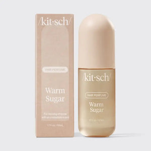 A bottle of KITSCH - HAIR PERFUME MIST IN WARM SUGAR stands between a slice of cake and blue cotton candy, with labeled fragrance notes: pink grapefruit, whipped cream, spun sugar, bergamot blossom, musk, vanilla, and praline. This odor-eliminating formula from KITSCH ensures your hair smells irresistibly sweet and fresh all day.