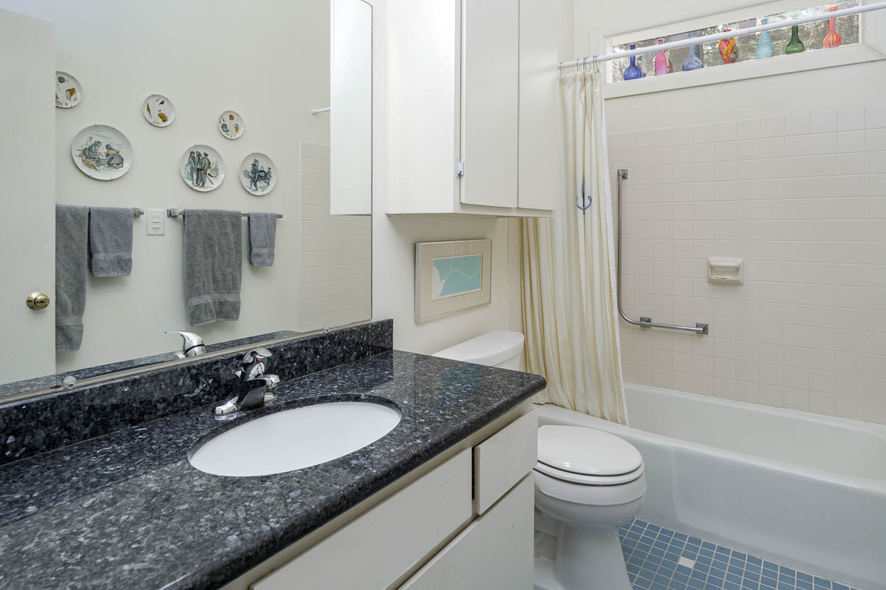 A clean bathroom with a black granite countertop, white sink, toilet, shower with a cream curtain, blue tile floor, wall art, gray towels, and colorful glass decor on a window shelf.