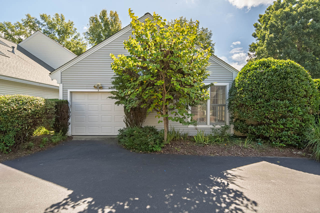 Single-story house with light gray siding, a white garage door, and a tree partially obscuring the front entrance. The driveway is paved and surrounded by green shrubs and trees.