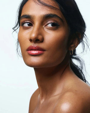 A woman with light skin and brown hair tied back wears small gold hoops and soft peach makeup. Her glossy pink lips, thanks to U BEAUTY’s THE PLASMA LIP COMPOUND IN BELLINI, catch the light as she gazes softly at the camera, a strand of hair falling across her face.