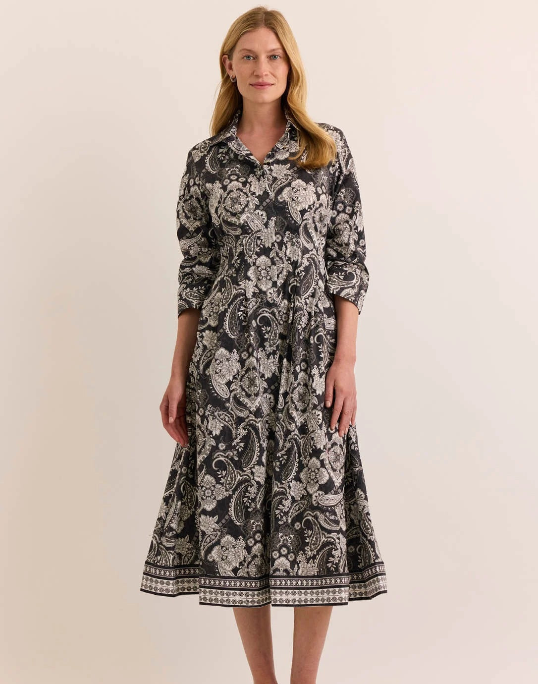 A woman stands wearing the HINSON WU - MARTINA 2/4 SLEEVE PAISLEY PRINT DRESS. This refined cotton dress by HINSON WU features a black background with a white floral and paisley print, a collared neckline, mid-calf length, flared skirt, side pockets, and elegant 2/4 sleeves.