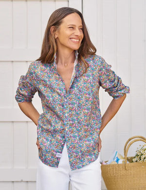 A woman with long brown hair smiles as she stands outside by a white wooden wall, wearing the FRANK & EILEEN EILEEN Relaxed Button Up in Italian Classic Linen Multicolor Flowers, paired with white pants and a straw tote bag nearby.