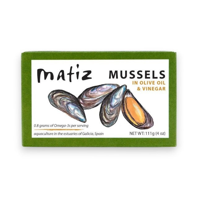 A green box of MATIZ ESPANA - MATIZ Mussels in Extra Virgin Olive Oil highlights its omega-3 content and origin from Galicia, Spain, with illustrations of Spanish mussels. Net weight: 111g.