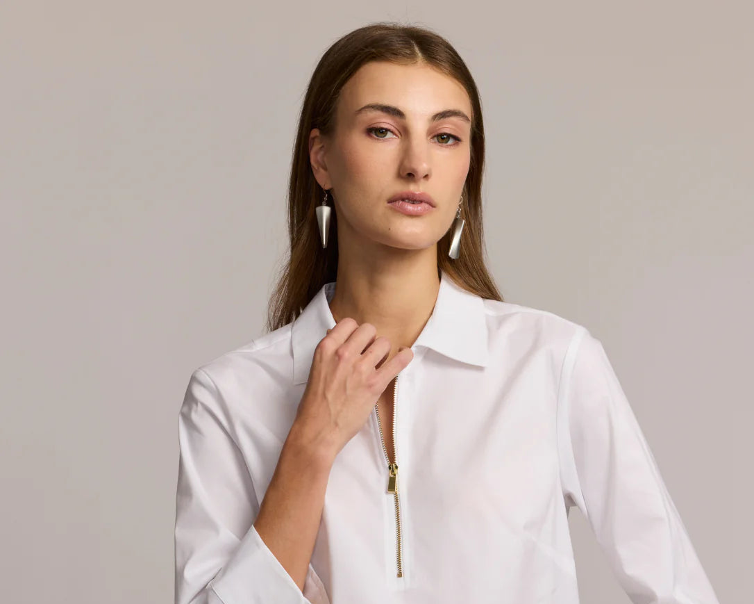 A woman with long brown hair wears a white collared shirt with a zippered front and white geometric earrings, looking confidently at the camera against a plain, light background.