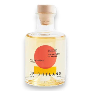A clear glass bottle of Brightland 'Parasol' Citrus Champagne Vinegar with a wooden stopper. The label features a bold design with an orange circle and yellow semicircle. Text on the label reads "Parasol Champagne Vinegar with California Citrus, Navel and Valencia oranges, Brightland, 200 mL, 6.7 oz.