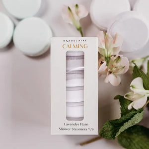 A BAUDELAIRE - CALM LAVENDER HAZE SHOWER STEAMER rests atop a slatted bamboo soap dish emblazoned with “BAUDELAIRE,” water droplets scattered on the white surface below—creating a tranquil scene evocative of soothing aromatherapy.