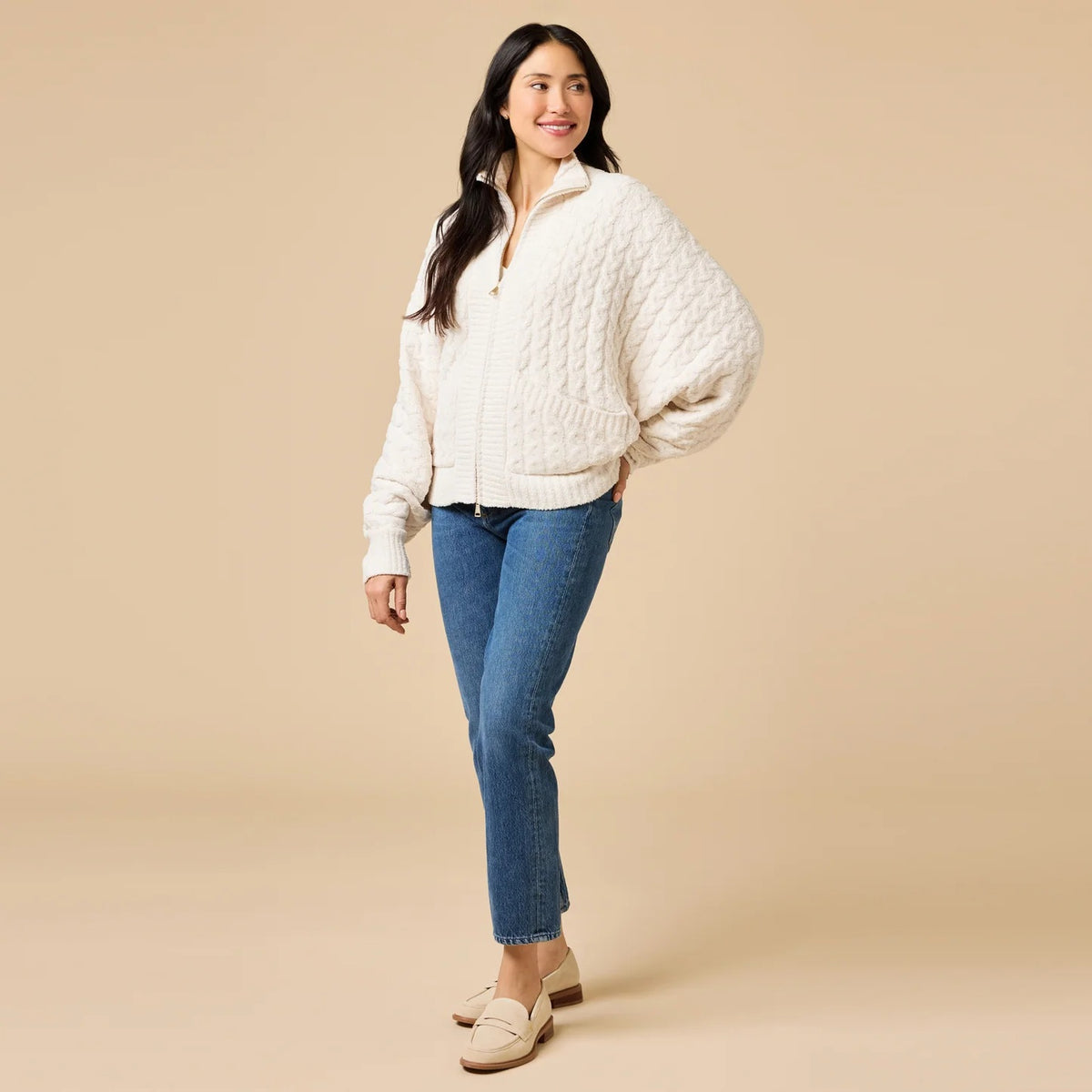 A woman wearing a cream-colored, plush SOFTIES - CABLE KNIT MARSHMALLOW FUNNEL NECK DOUBLE ZIP JACKET with a funnel neck and double zipper, paired with blue jeans.
