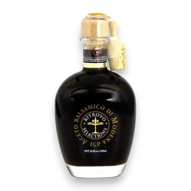 A clear glass bottle of RITROVO SELECTIONS - AGED BALSAMIC VINEGAR by RITROVO, an authentic Italian condiment, featuring a cork stopper and a black and gold label, displayed against a white background.
