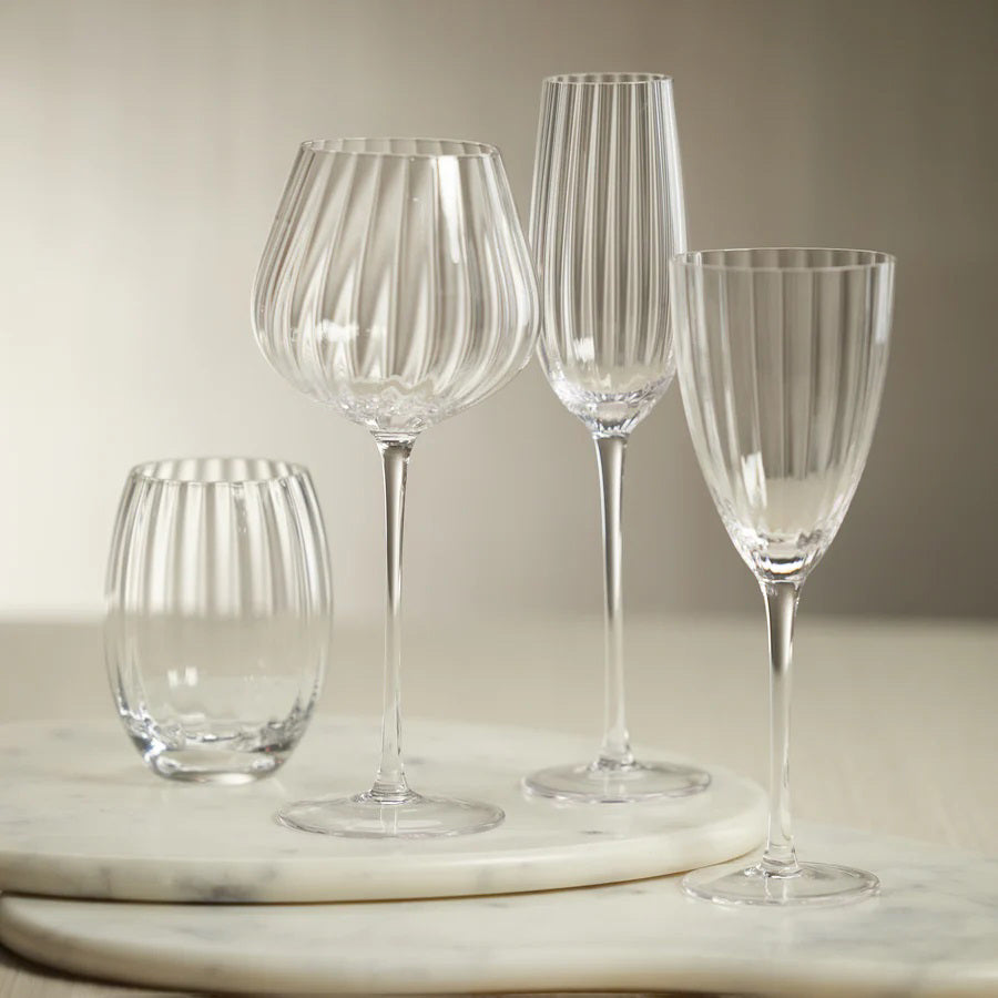 A MADELEINE OPTIC RED WINE GLASS by ZODAX stands on a white background. This elegant glass, with a capacity of 580 ml, features a long, slender stem and a wide, slightly curved bowl adorned with vertical ribbed detailing. Its round, flat base ensures stability for the delicate glassware.