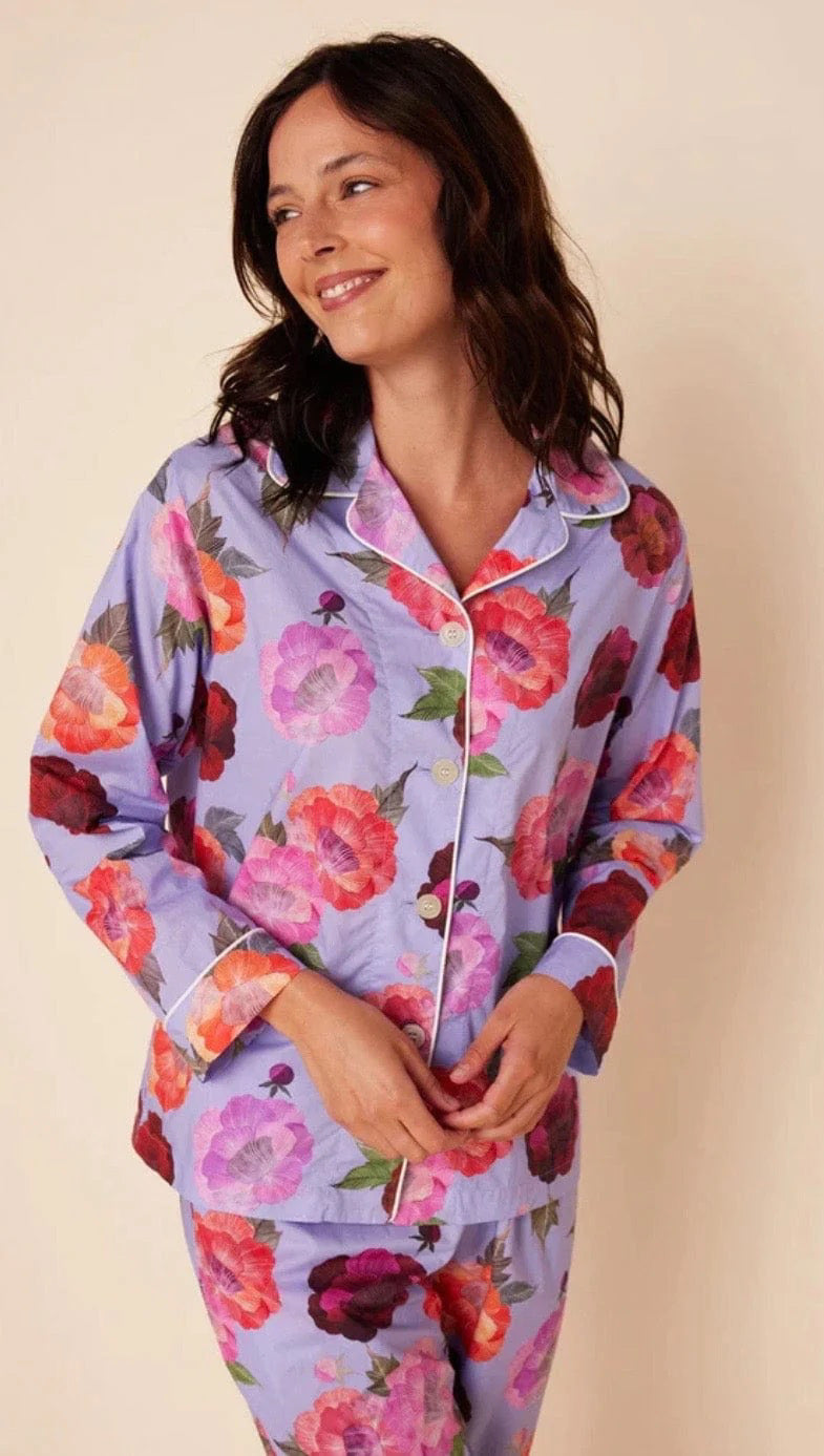 A woman is standing against a plain background, smiling downwards with her hands in the pockets of her Cat&#39;s Pajamas - Lavender Floral Luxe Pima Cotton Pajamas. The pajama set from Cat&#39;s Pajamas features a vibrant floral pattern with large red, pink, and purple flowers. She is barefoot and appears relaxed.