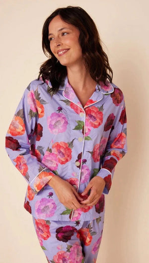 A woman is standing against a plain background, smiling downwards with her hands in the pockets of her Cat's Pajamas - Lavender Floral Luxe Pima Cotton Pajamas. The pajama set from Cat's Pajamas features a vibrant floral pattern with large red, pink, and purple flowers. She is barefoot and appears relaxed.