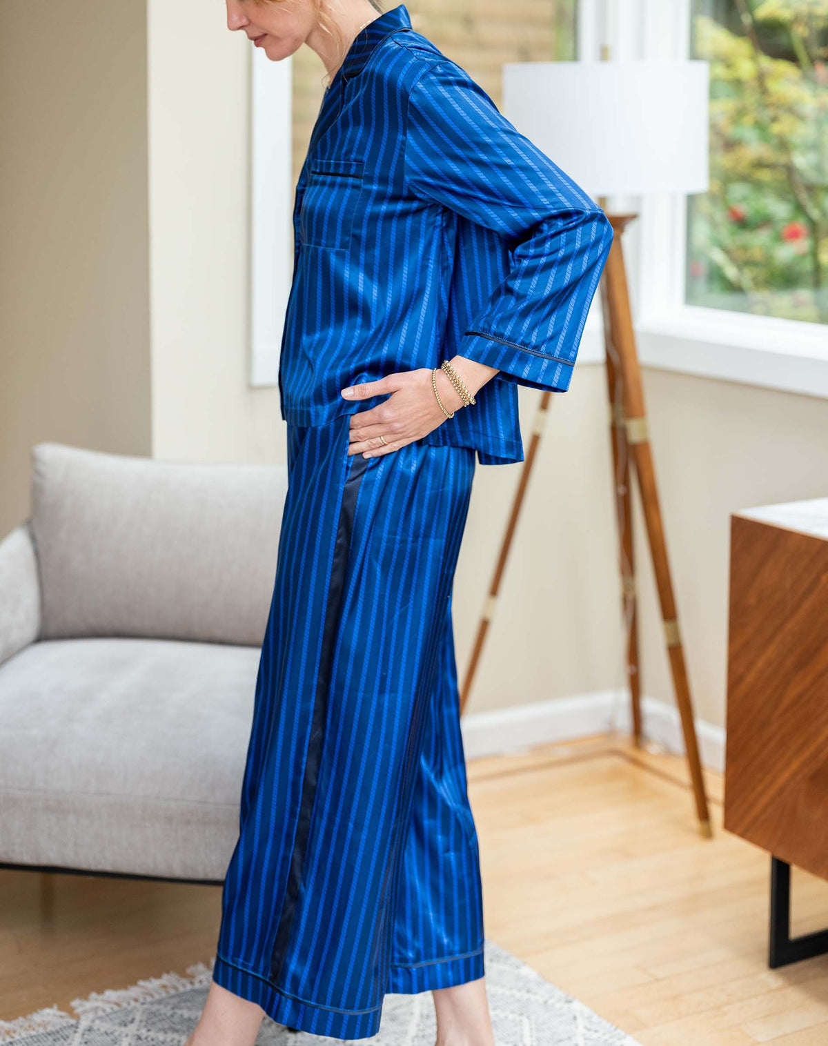 The MERSEA - Carousel Stripe Satin Sailor Pajama Set in Blue by MERSEA, featuring elegant blue and black stripes with a button-up top and matching pants, embodies luxury sleepwear and is neatly folded on a light blue background.