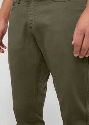 A person stands facing away from the camera against a plain background, wearing DUER - NO SWEAT ATHLETIC TAPER PANTS IN KELP paired with brown shoes.