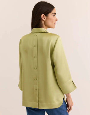 A woman with long brown hair is wearing the HINSON WU - AILEEN 3/4 Sleeve Liquid Satin Shirt in Pistachio, a light green collared blouse with three-quarter sleeves, paired with blue jeans as she stands against a plain, light-colored background. The shirt by HINSON WU features an elegant drape and luminous sheen, adding a touch of sophistication to her relaxed look.