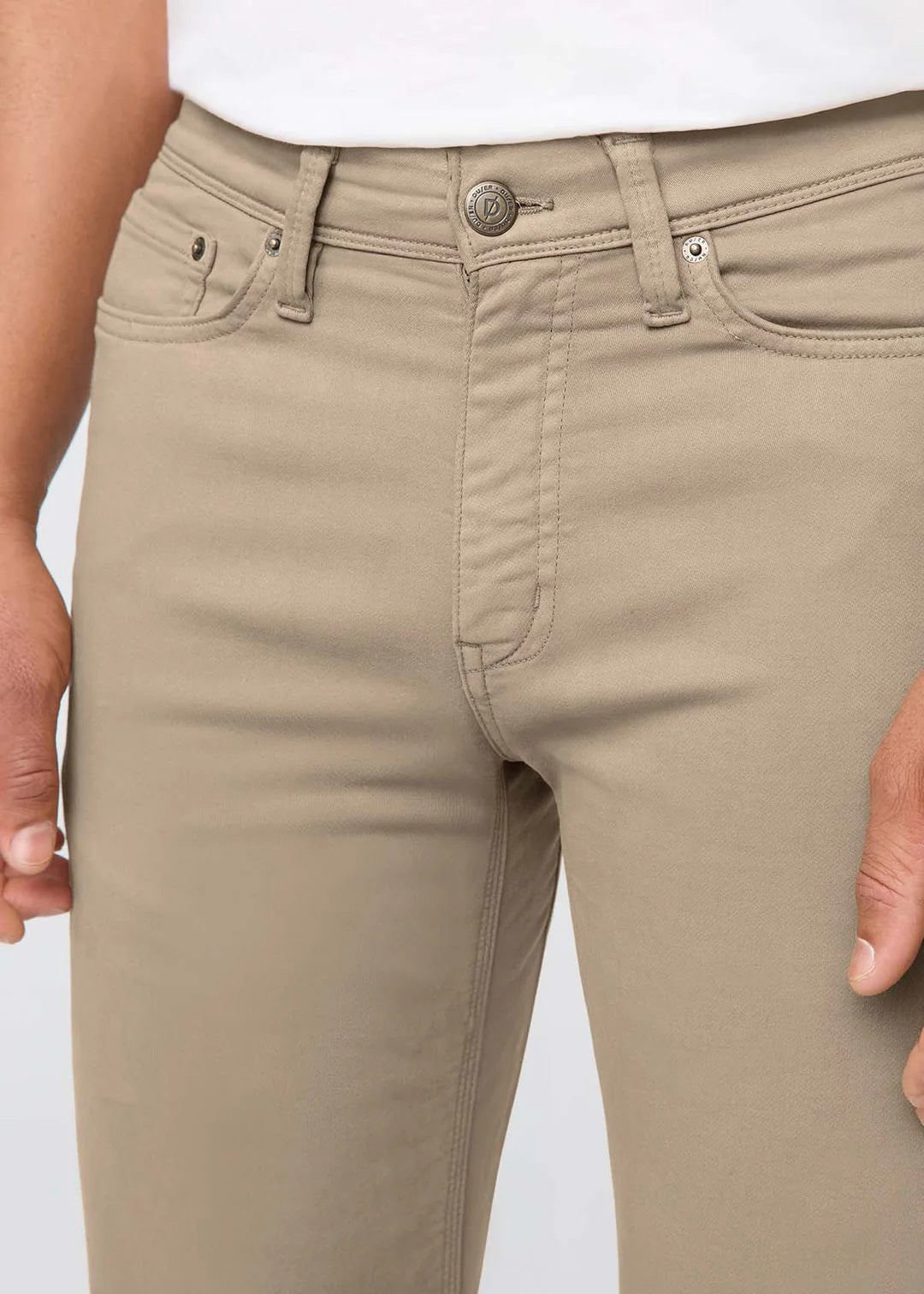 A person wearing DUER NO SWEAT STRAIGHT PANTS IN DESERT KHAKI, a white t-shirt, and white sneakers stands against a plain light gray background.