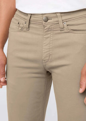 A person wearing DUER NO SWEAT STRAIGHT PANTS IN DESERT KHAKI, a white t-shirt, and white sneakers stands against a plain light gray background.