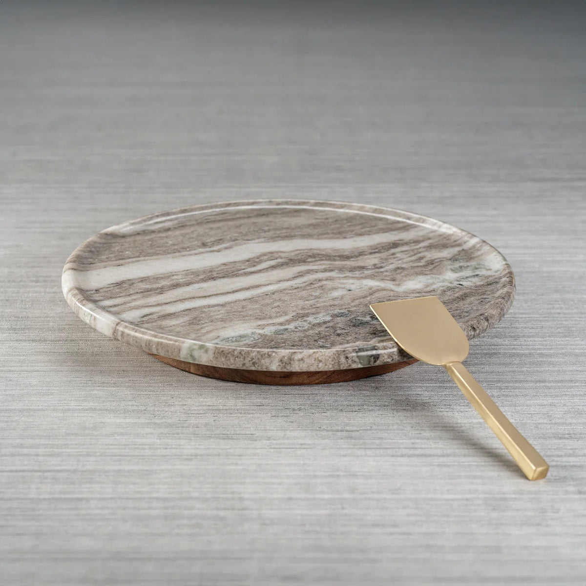 The ZODAX CREMA MARFIL MARBLE LAZY SUSAN, a round 16-inch grey and white serving platter, sits on a textured grey surface with a gold-colored metal spatula resting at its edge.