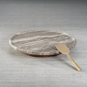The ZODAX CREMA MARFIL MARBLE LAZY SUSAN, a round 16-inch grey and white serving platter, sits on a textured grey surface with a gold-colored metal spatula resting at its edge.