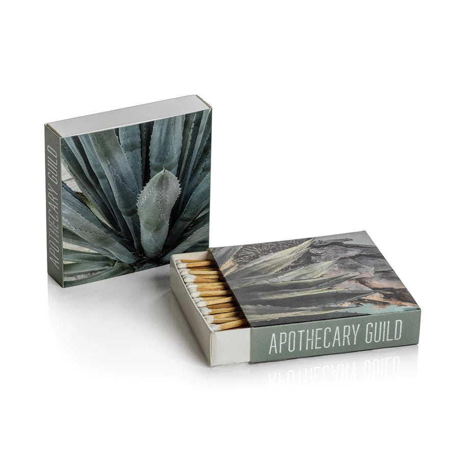 The MATCH BOX - AGAVE by ZODAX features two square designs—one closed and one open to reveal long, decorative matchsticks—with "ZODAX" branding on both the front and side.