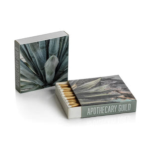 The MATCH BOX - AGAVE by ZODAX features two square designs—one closed and one open to reveal long, decorative matchsticks—with "ZODAX" branding on both the front and side.