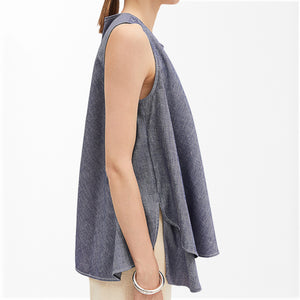A model wears the MEIMEJ - SLEEVELESS DENIM TUNIC by MEIMEIJ, a tunic-length, light cotton denim piece with side slits, styled with light-colored pants and a silver bracelet, shown in profile against a plain white background.