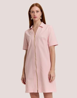 A woman with long, straight auburn hair wears the HINSON WU Bridget Short Sleeve Traveler's Twill Dress in blush pink, featuring a collar and gold front zipper, standing against a plain light background.