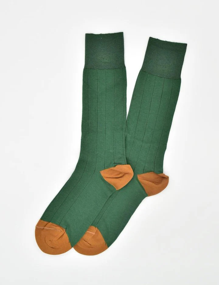A person in light pants and a purple checkered shirt pulls up BIRD DOG BAY - PEDIGREE MID-CALF SOCKS IN GREEN by BIRD DOG BAY against a light teal background.
