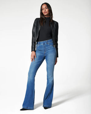 A woman poses against a white background in the SPANX jeannish high-waisted blue flare jeans made from premium stretch denim with hidden core shaping technology, styled with a black faux leather jacket, black top, and pointed-toe shoes.