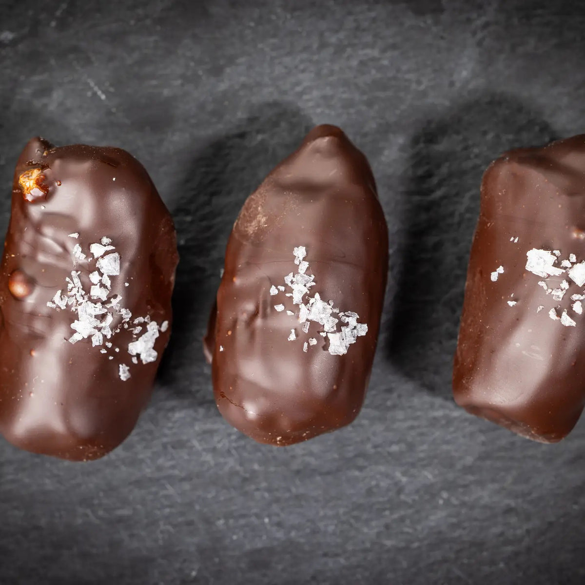 Three DATE BETTER - ALMOND JAVA CRUNCH COVERED DATES, by DATE BETTER SNACKS, arranged on a dark slate surface offer a deliciously healthy candy alternative.