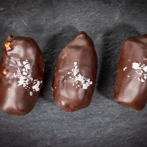 Three DATE BETTER - ALMOND JAVA CRUNCH COVERED DATES, by DATE BETTER SNACKS, arranged on a dark slate surface offer a deliciously healthy candy alternative.