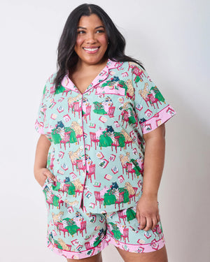 Dressed in PRINTFRESH’s Mahjong Menagerie Pajama Short Set, this person sports matching pastel organic cotton sleepwear featuring a playful cat print with tables, books, and chairs. The set includes a short-sleeve button-up shirt and shorts, both finished with light pink trim.