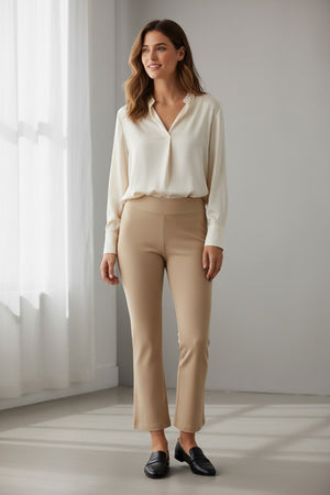 A woman stands indoors by a large window, wearing a long-sleeve cream blouse and the KRAZY LARRY, INC PULL ON WIDE ANKLE PANT in black, which features a tummy tuck design. She completes her look with black loafers and smiles slightly, her wavy brown hair catching the sunlight that casts shadows on the white floor and wall.